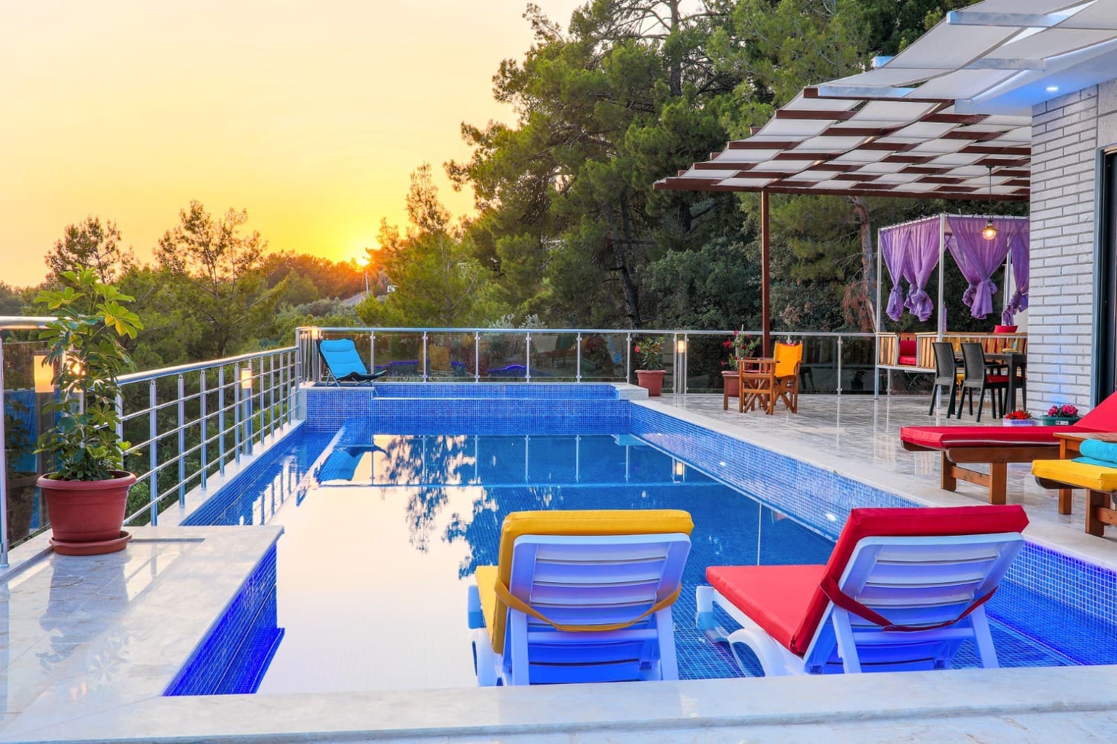 Gün batımında havuz başında renkli minderli şezlonglar ve ağaçlar görünmekte.