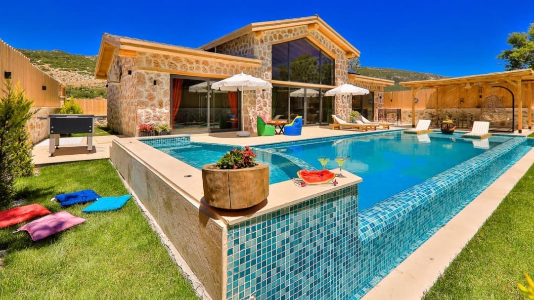 Taş cepheli iki katlı villanın önünde yeşil çim alan ve masmavi havuz.