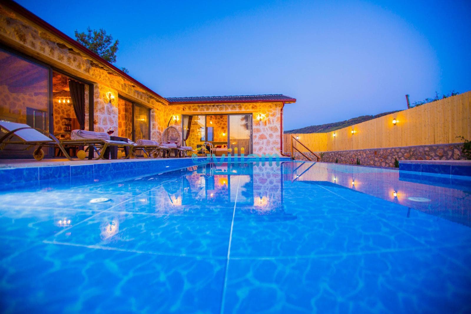 Akşam saatlerinde aydınlatılmış taş villa ve önünde parlayan yüzme havuzu görünüyor.