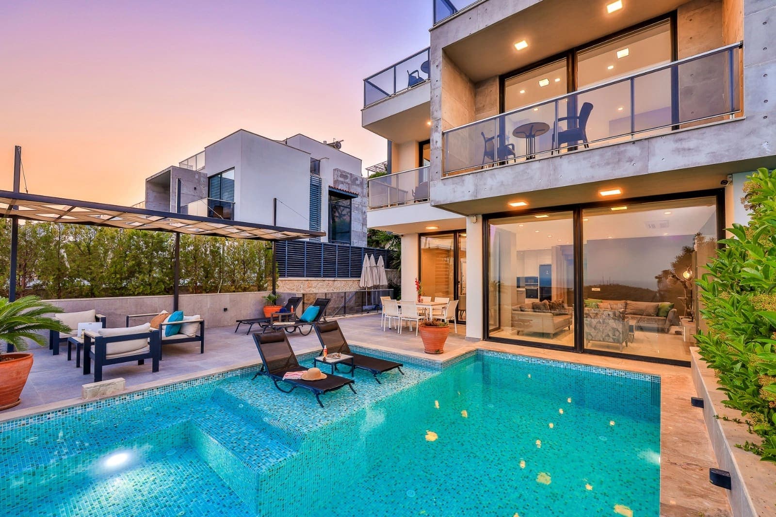 Akşam üstü ışıklandırılmış havuz ve oturma grubu olan modern villa dış cephesi.