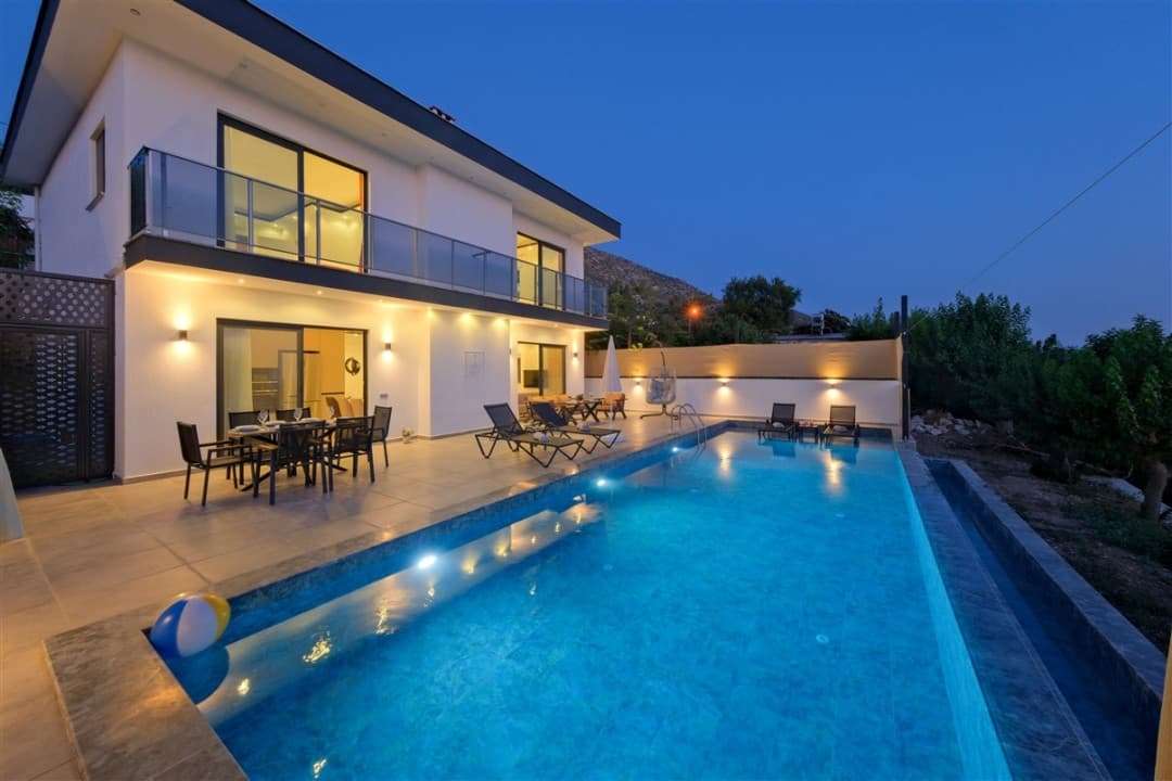 Akşam ışıklandırmalı, havuzlu ve terasında yemek masası ile şezlonglar bulunan iki katlı modern villa.