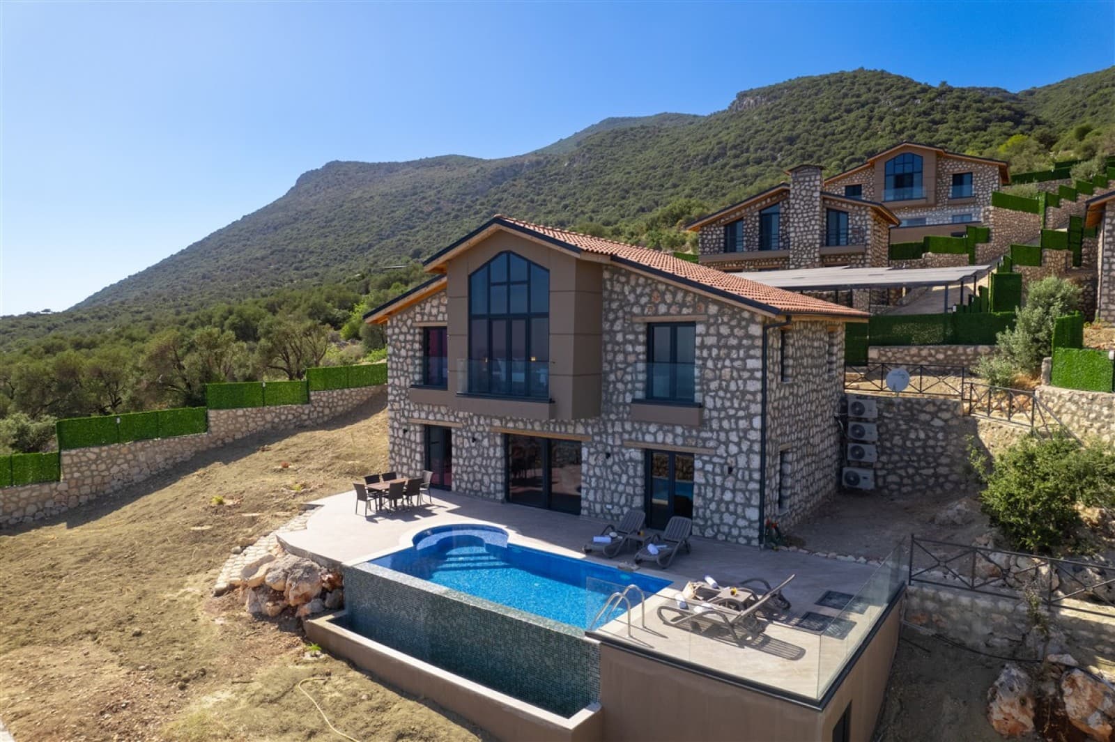 Dağ etek kurulu taş cepheli villa, önünde havuz ve şezlonglar.