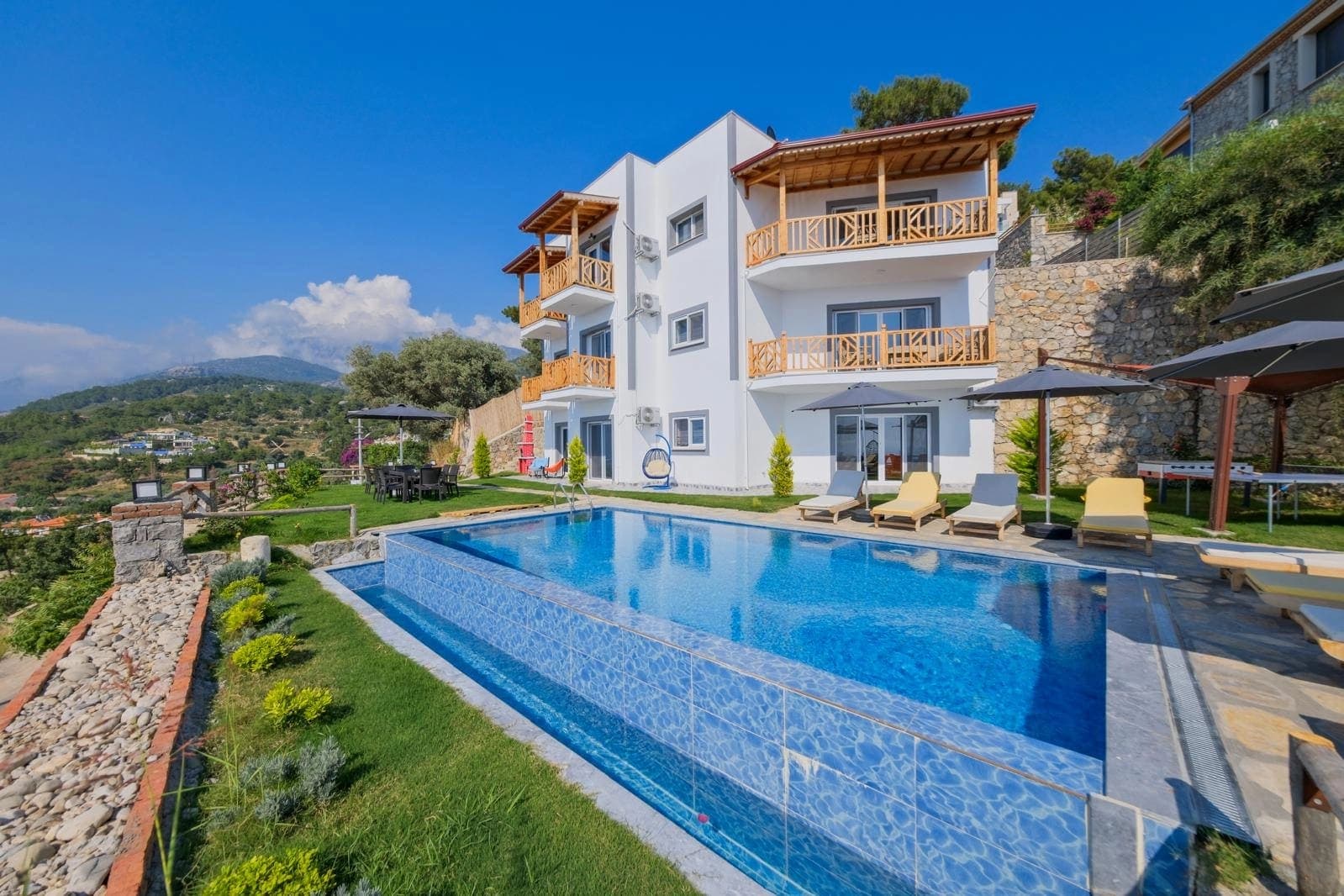 Önünde havuzu, şezlongları ve ahşap detaylı balkonları bulunan beyaz villanın dış görünümü.