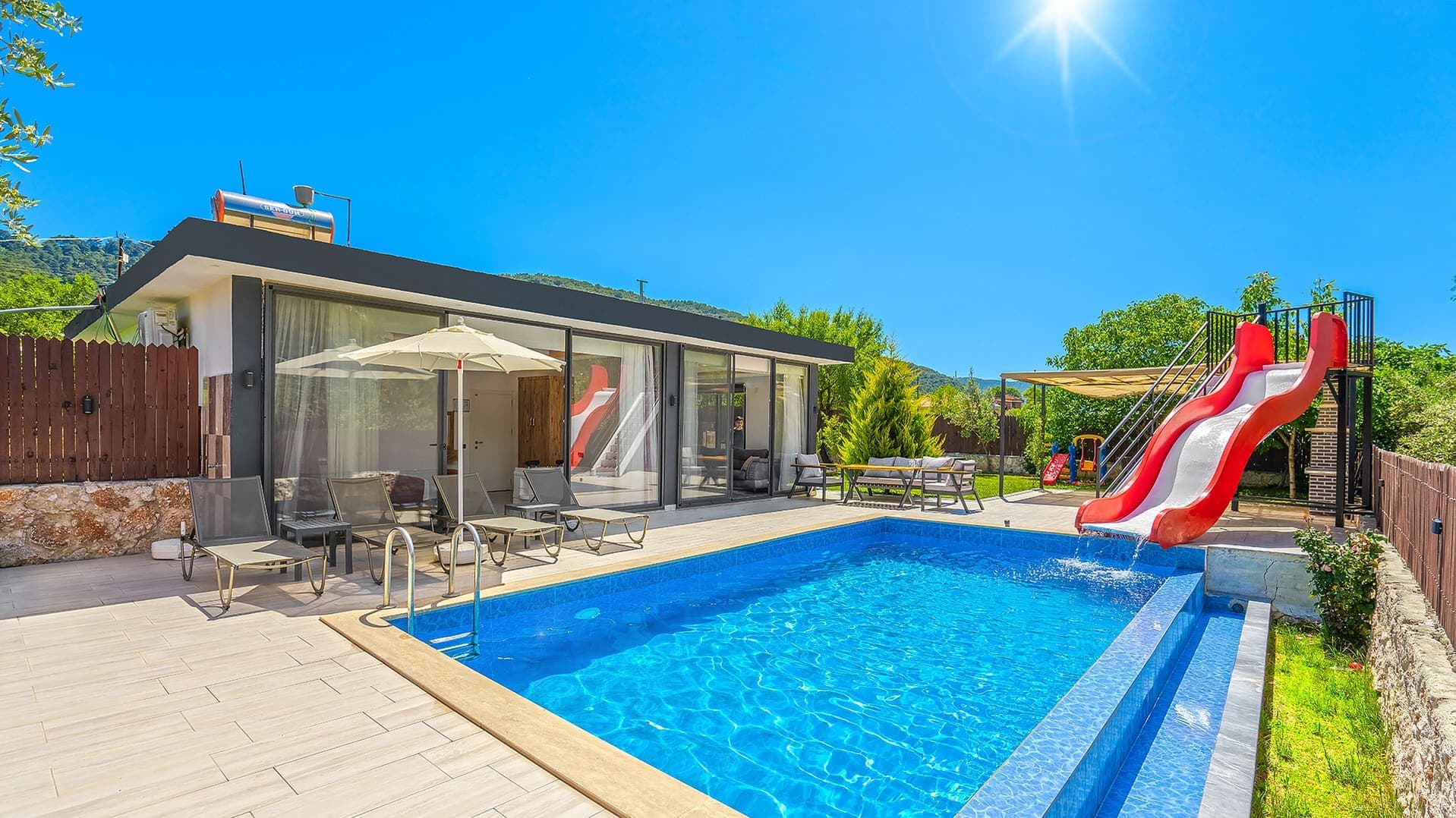 Havuz kenarında kırmızı kaydırak, şezlonglar ve güneş şemsiyesi bulunan tek katlı villa.
