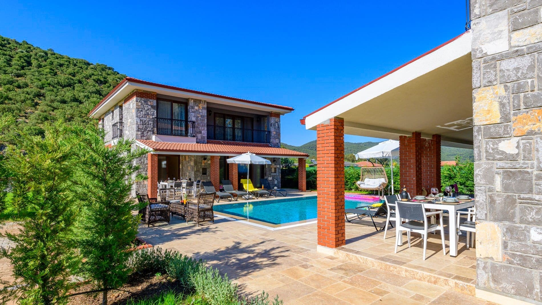 İki katlı taş villa ve önünde açık yüzme havuzu ile şezlonglar.