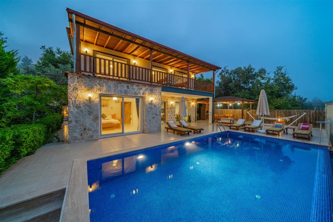 Akşam vakti aydınlatılmış, taş duvarlı, havuzlu ve balkonlu iki katlı villa.
