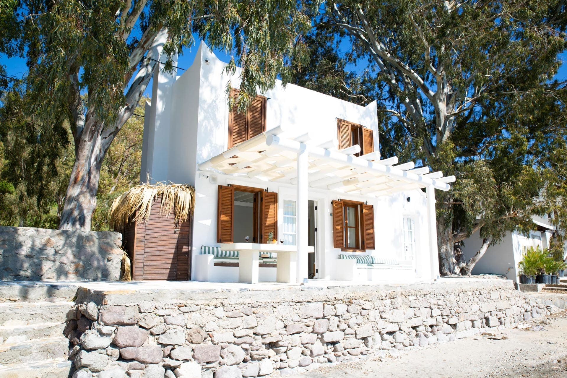 Ağaçlar arasında yer alan, kahverengi panjurlu beyaz dış cepheli iki katlı villa.