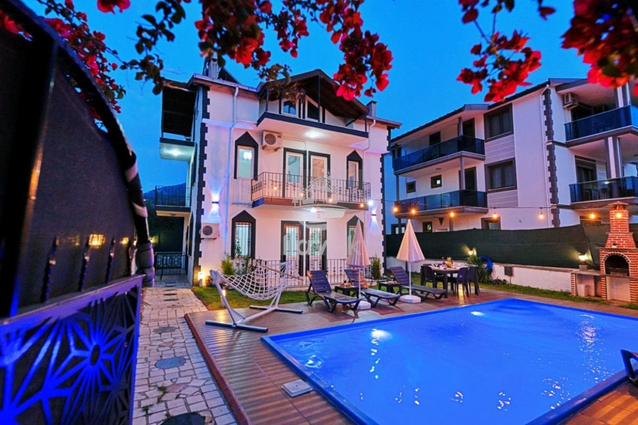 Akşam saatlerinde havuz başı şezlongları ve aydınlatmalı dış cephesiyle villa.