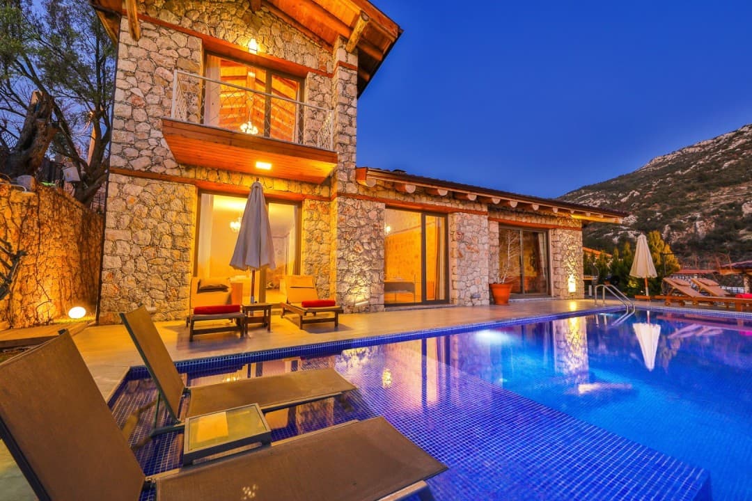 Akşam vakti aydınlatılmış taş duvarlı villa, önünde yüzme havuzu ve şezlonglar bulunuyor.