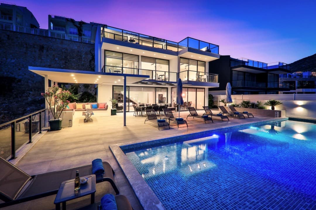 Aydınlatılmış geniş havuz kenarında şezlonglar ve gün batımında modern villa dış cephesi.