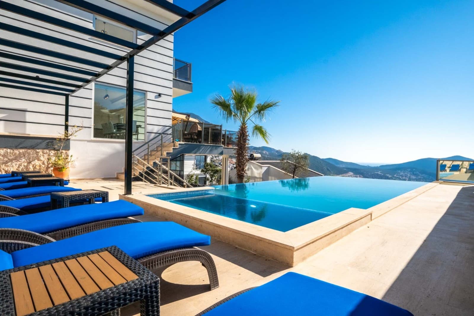 Ahşap şezlonglu havuz başı terası, beyaz dış cepheli villa ve dağ manzarası.