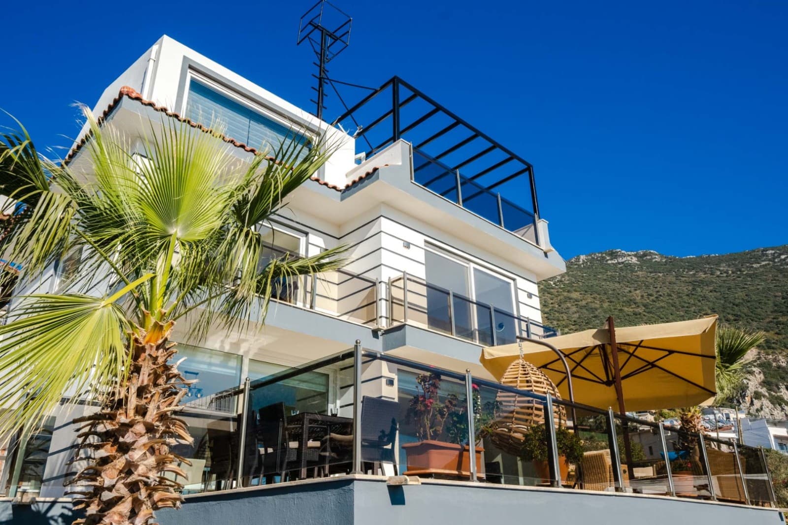 Mavi gökyüzü altında palmiye ve şezlonglu villa dış cephesi.