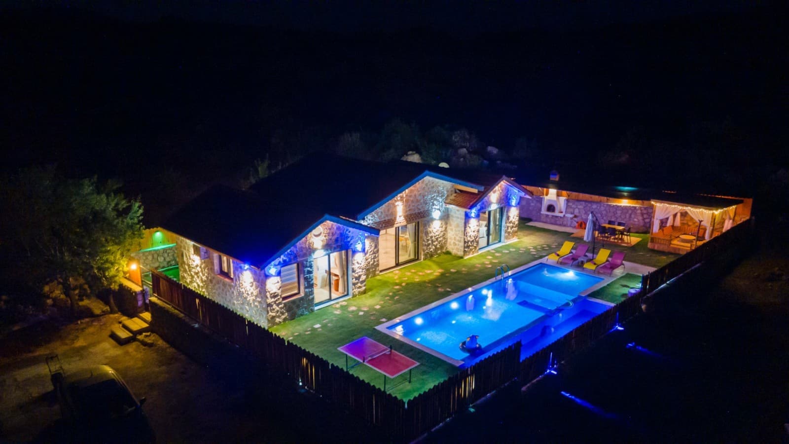 Gece aydınlatılmış taş villa, havuz, çardak ve masa tenisi bulunan bahçe.