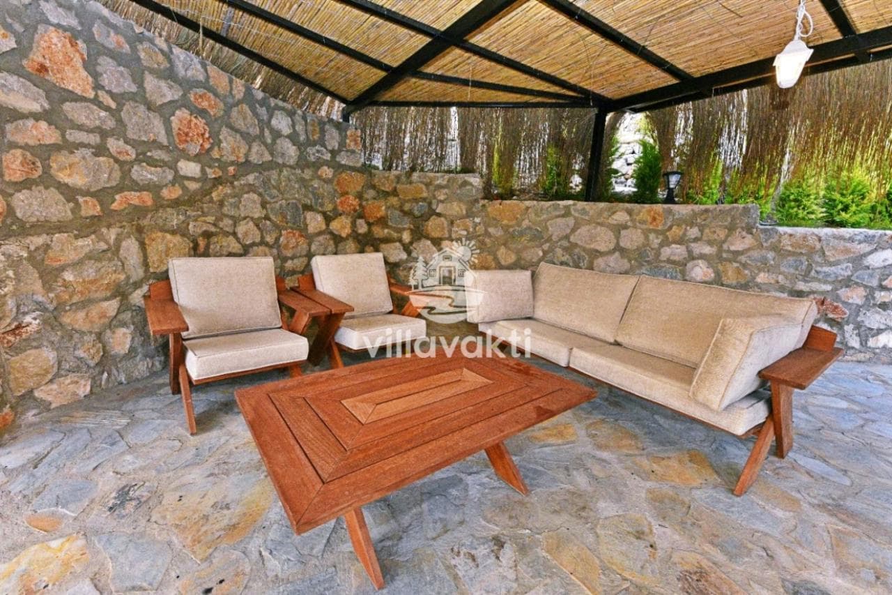 Taş duvarlı verandada ahşap sehpa ve bej renkli minderlere sahip koltuklar yer alıyor.