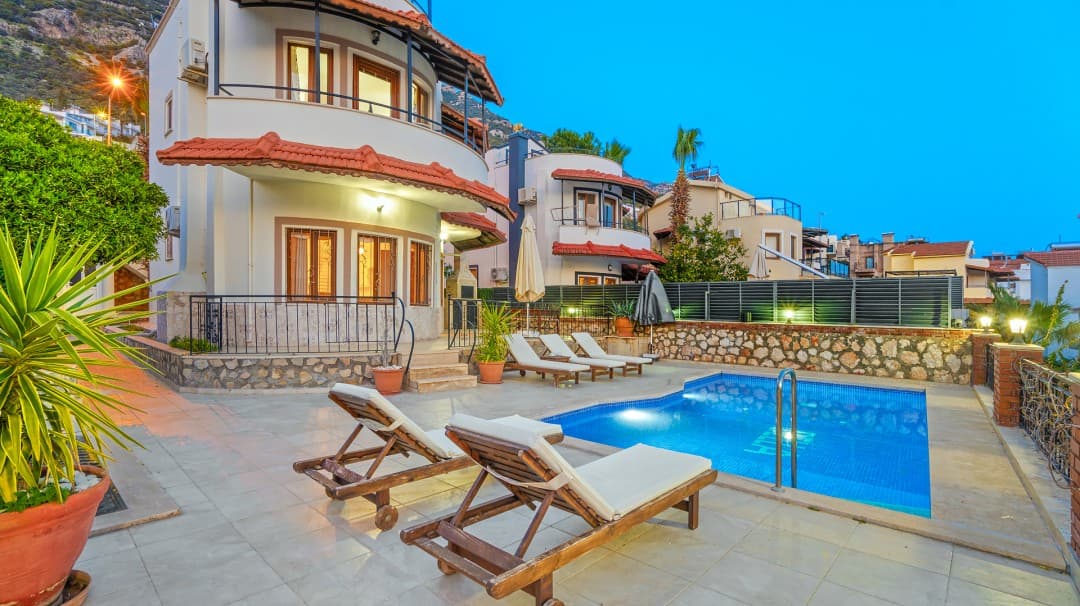 Açık havuzun başında ahşap şezlonglar ve arkada iki katlı villa yer alıyor.