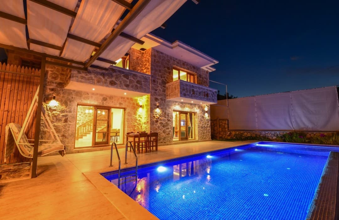 Gece aydınlatmalı taş villa, önünde havuz, ahşap salıncak ve dış mekan yemek masası bulunuyor.