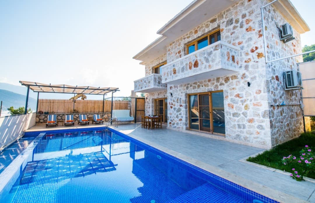 Taş kaplama iki katlı villanın önünde mavi havuz ve ahşap şezlonglar var.