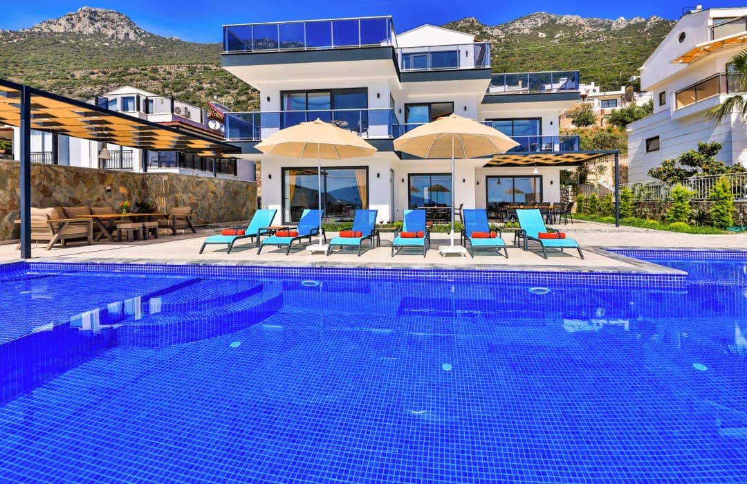 Ön planda mavi mozaikli havuz, havuz kenarında şezlonglar, şemsiyeler ve arkada beyaz katlı villa.