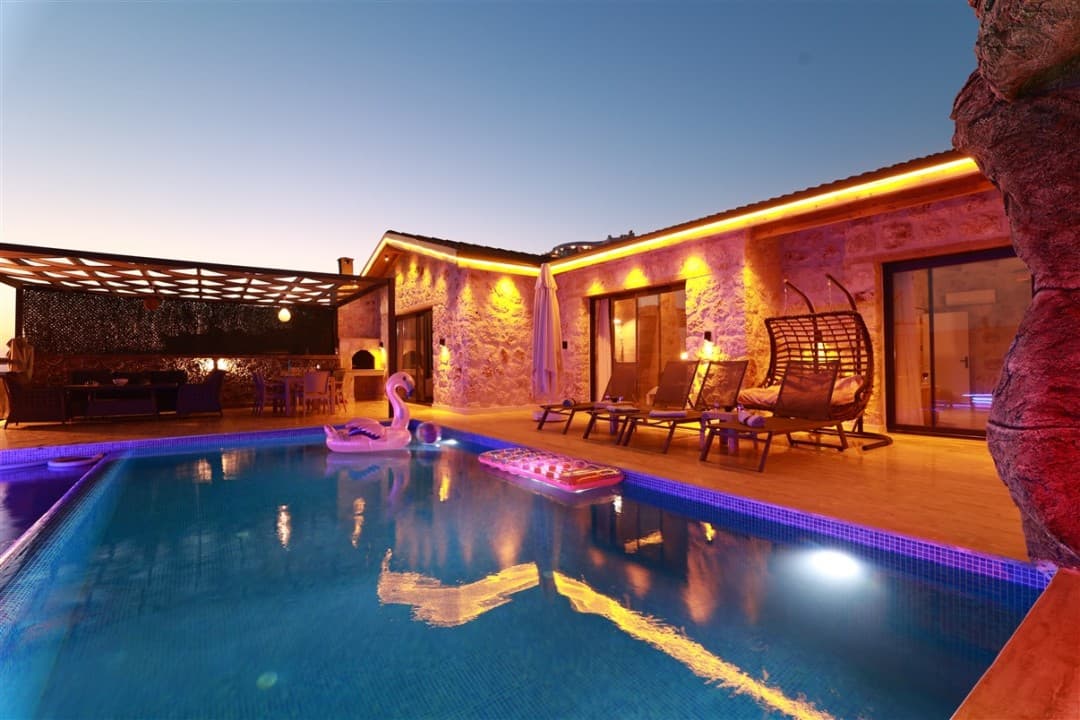 Akşam vakti aydınlatılmış taş villa, özel havuz, şezlonglar, salıncak ve havuz üzerinde flamingo deniz yatağı.