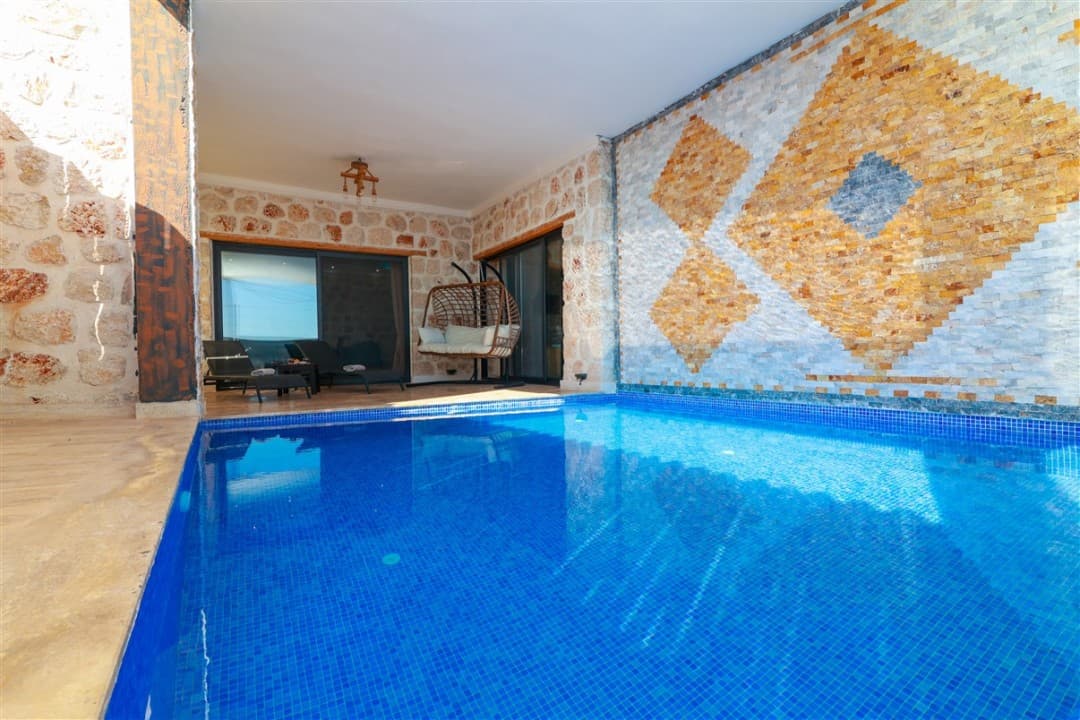 Taş duvarlı, kapalı havuzlu terasta asılı salıncak ve şezlonglar bulunan modern villa bölümü.