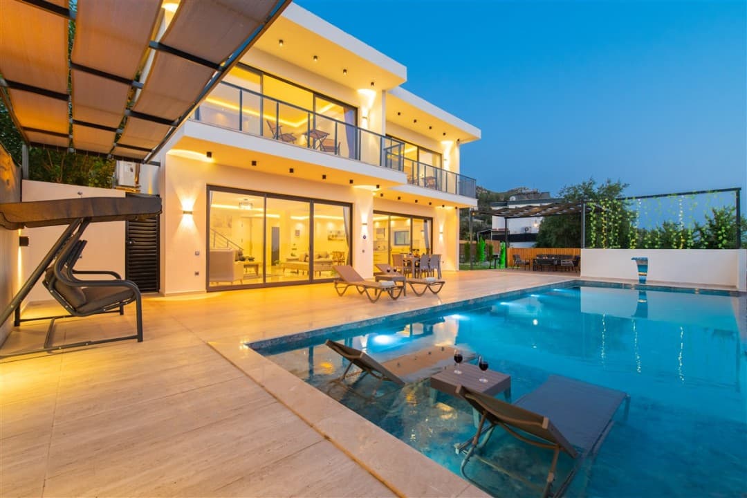 Akşam ışıklandırmalı, havuz içi şezlongları ve bahçe salıncağı bulunan iki katlı modern villa.