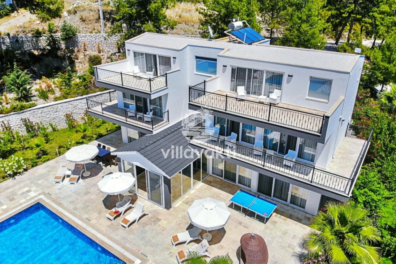 Ağaçlık yamaca kurulmuş, çok katlı ve balkonlu beyaz villanın havuz ve teras alanı.