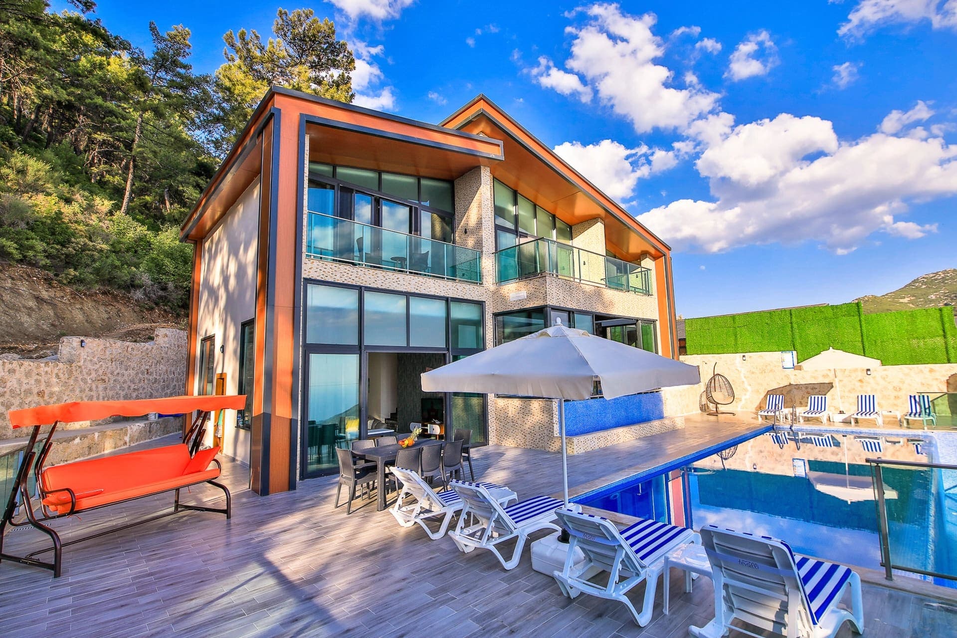 Ahşap ve taş detaylı modern villanın önünde havuz, şezlonglar ve salıncak bulunuyor.