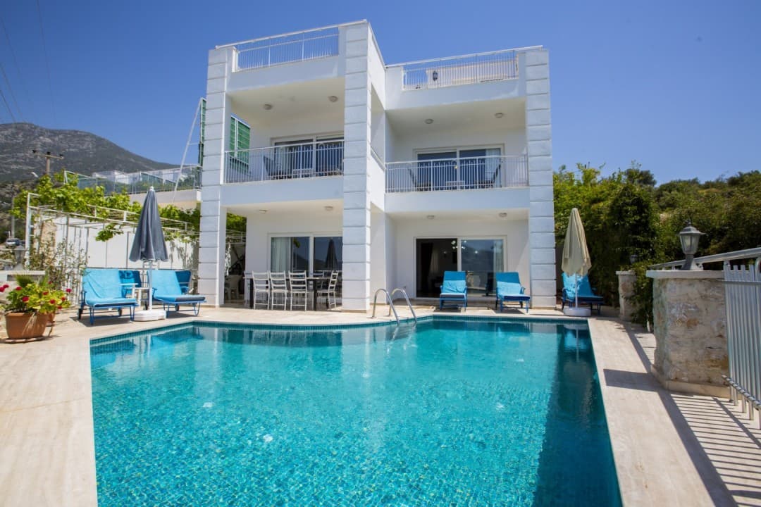 Mavi şezlonglu havuzun arkasında iki katlı beyaz villa ve dağ görünmektedir.