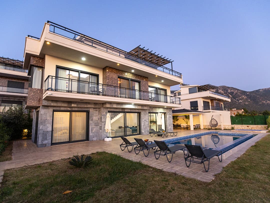 Gün batımı ışığında taş kaplama detaylı, havuzlu ve şezlonglu modern villa dış görünümü.