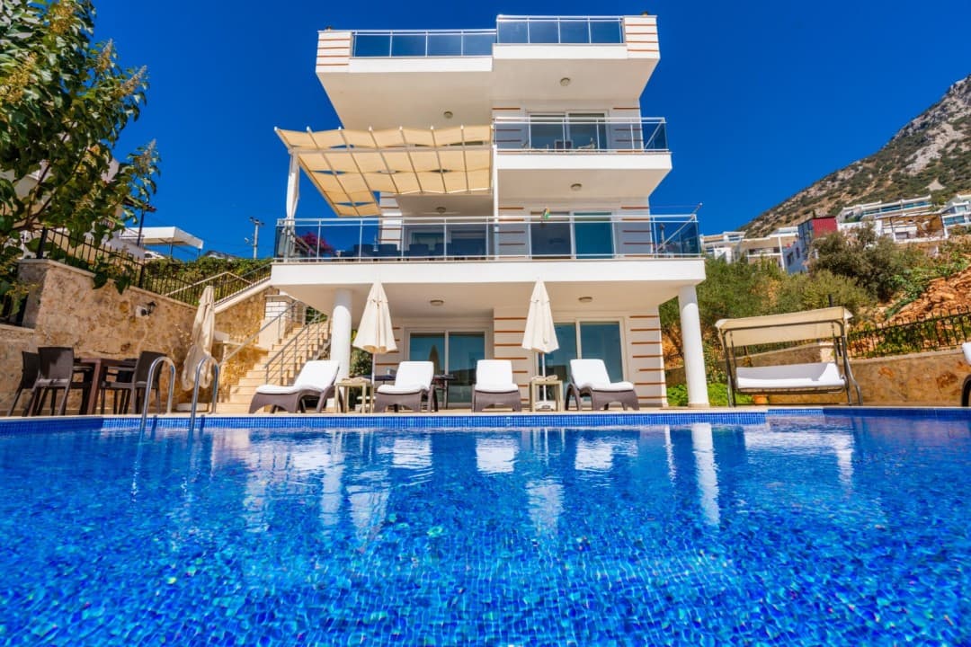Açık mavi havuzun arkasında beyaz dış cepheli üç katlı villa ve şezlonglar yer alıyor.