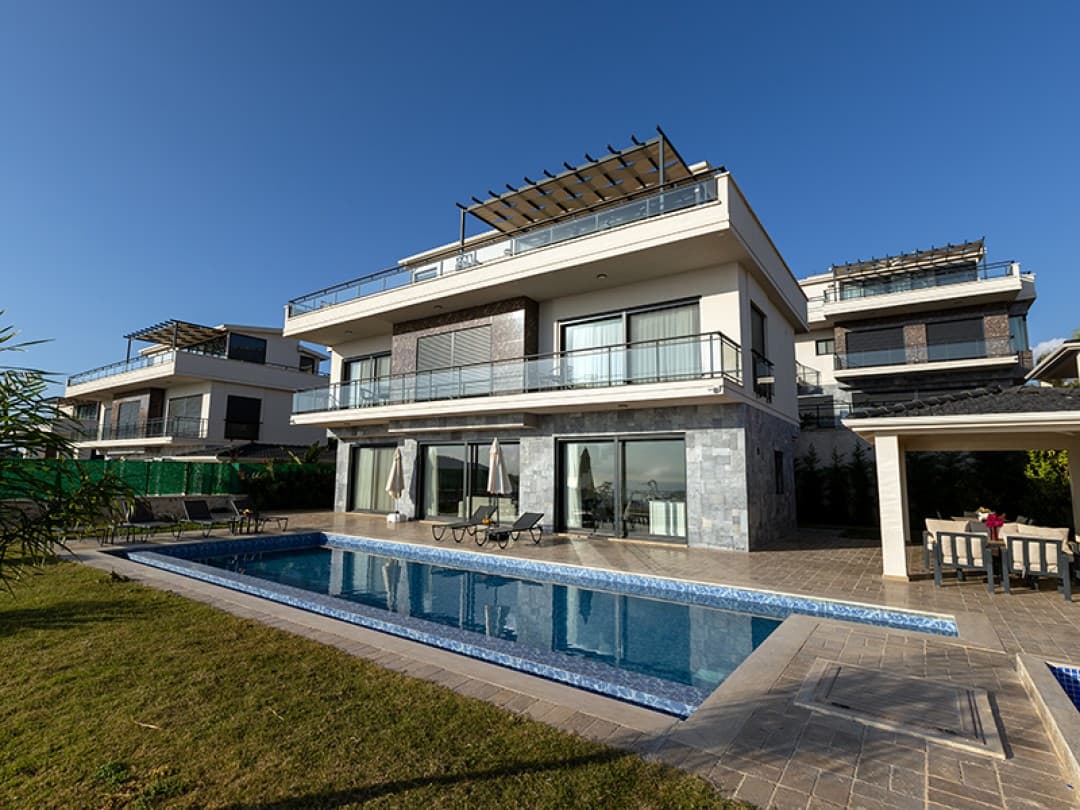 Mavi gökyüzü altında, önünde yüzme havuzu ve şezlonglar bulunan modern taş kaplamalı üç katlı villa.