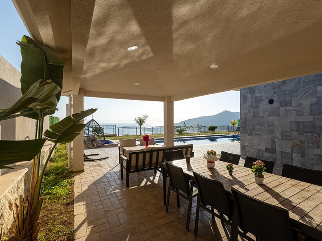 Geniş verandada ahşap yemek masası ve oturma grubu, arkada havuz ve deniz manzarası.