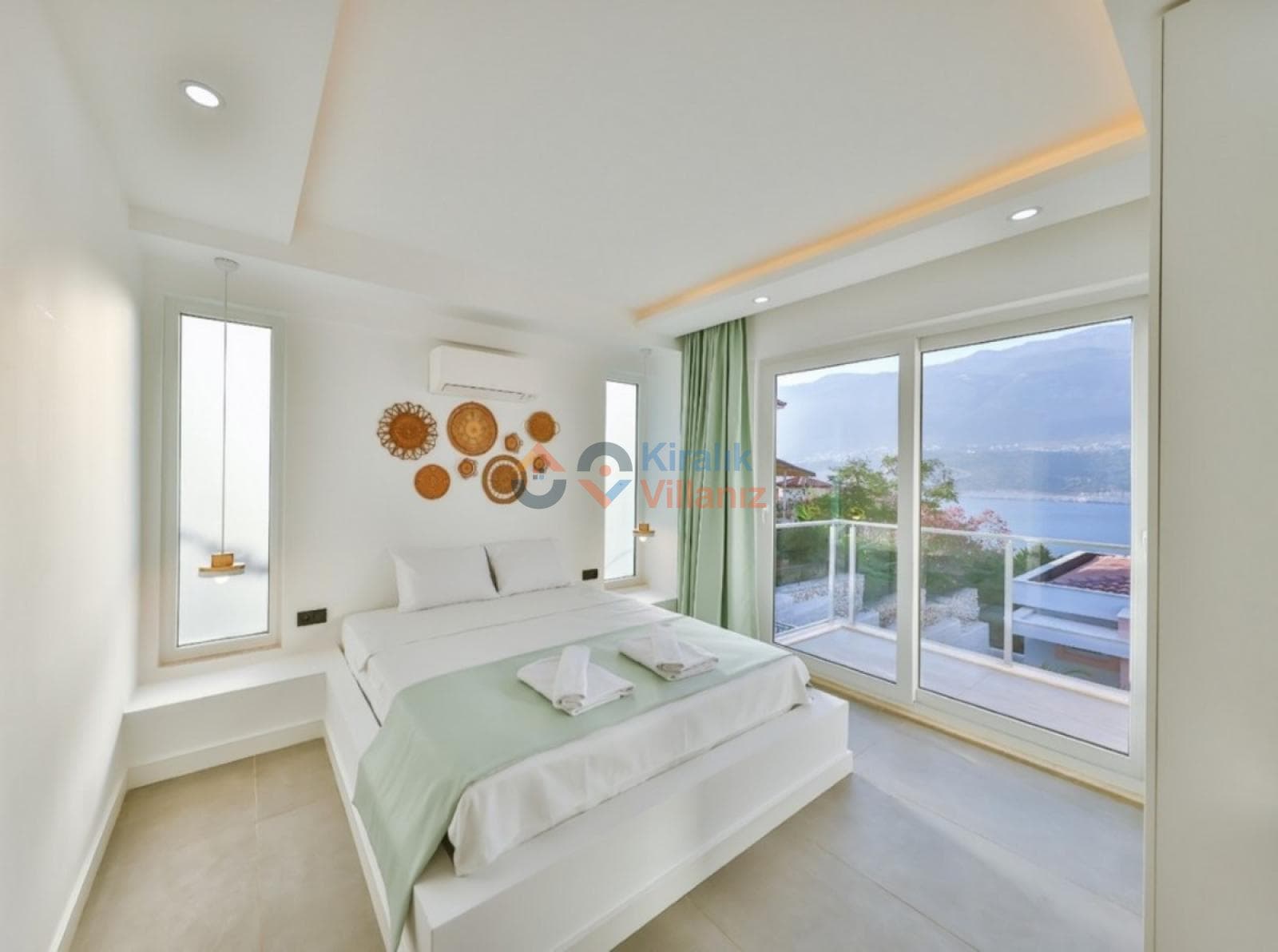 Beyaz ağırlıklı yatak odasında çift kişilik yatak, duvar süsleri ve deniz manzaralı geniş balkon kapısı var.