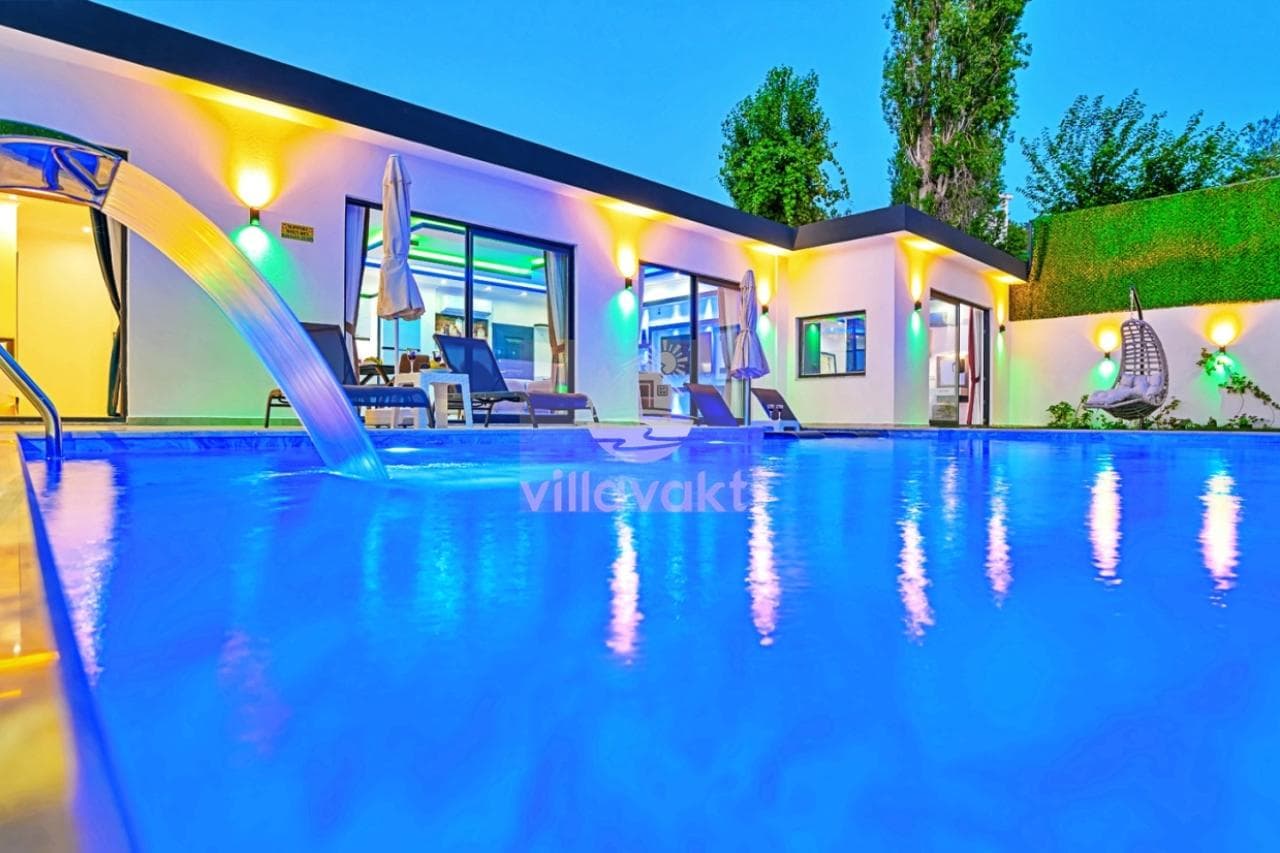 Akşam ışıklandırılmış modern villanın önünde mavi havuz ve şezlonglar bulunuyor.