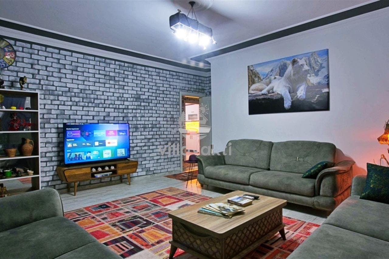 Gri tuğla duvarlı oturma odasında gri koltuklar, televizyon ve sehpa bulunuyor.