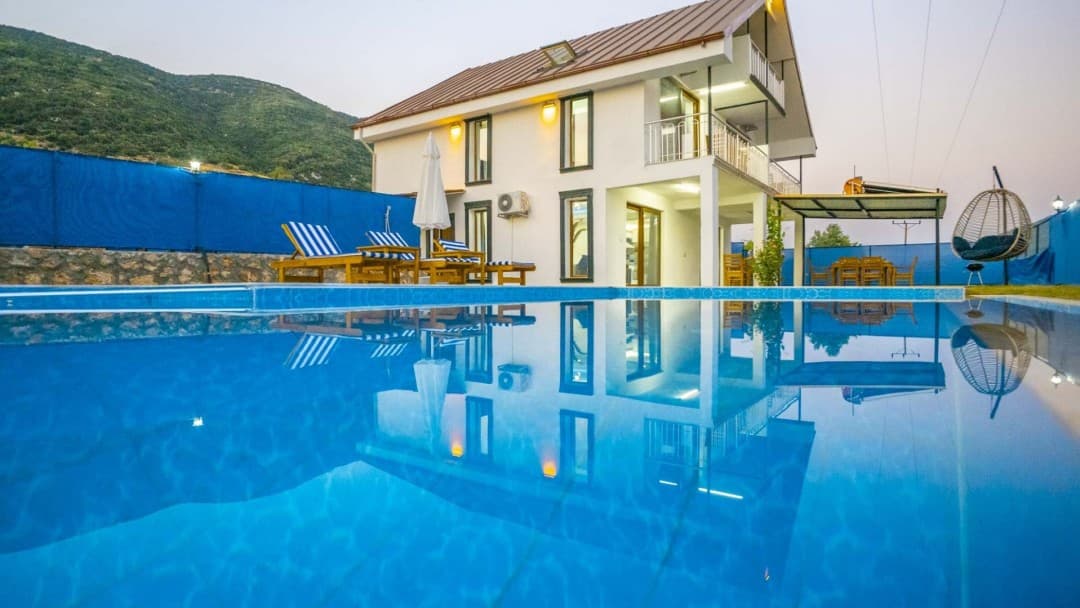 Ön planda havuz, arka planda şezlonglar, salıncak ve dağ manzaralı iki katlı beyaz bir villa bulunuyor.