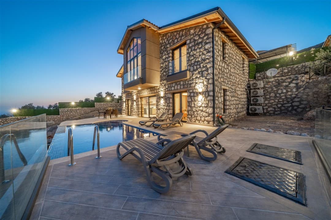 Akşam saatlerinde taş duvarlı, aydınlatmalı villa ve önündeki havuz alanı.