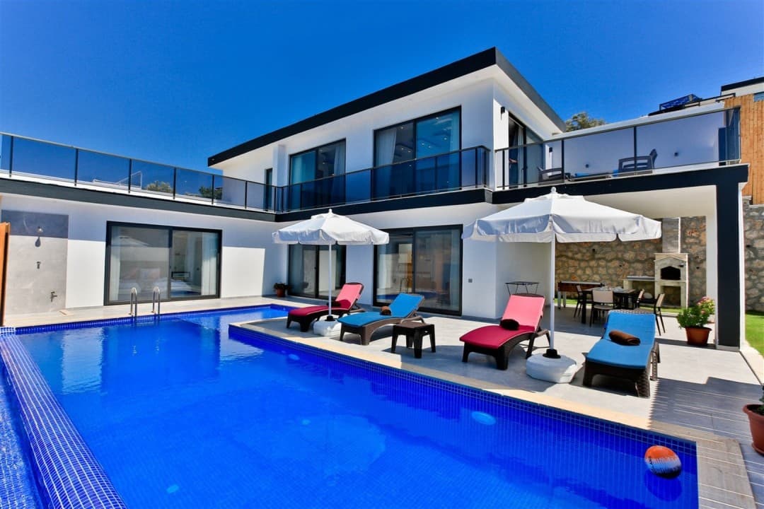 Gündüz vakti havuz kenarında şezlonglar ve arka planda beyaz villa.