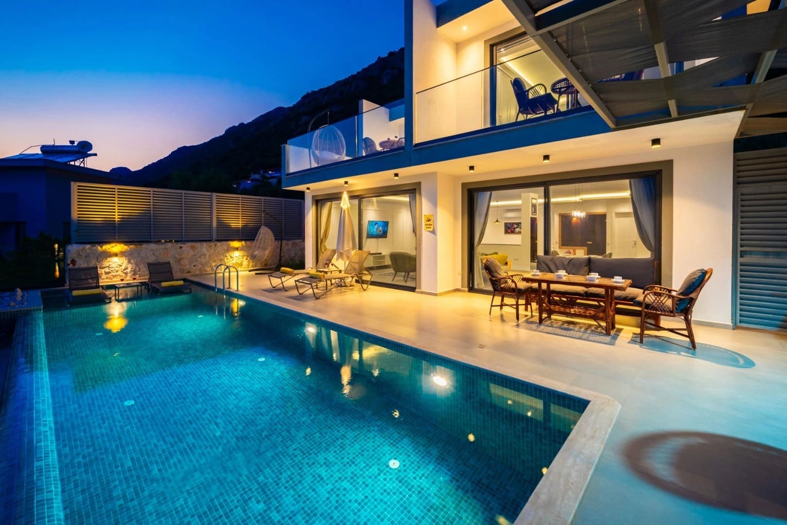 Aydınlatmalı havuz kenarında masa ve sandalyeler bulunan villa dış cephesi.