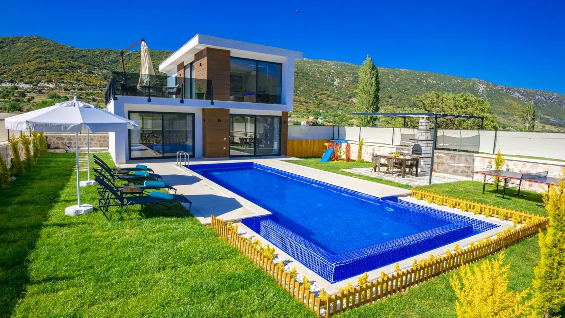 Bahçesinde havuz, şezlonglar ve masa bulunan iki katlı modern villa.