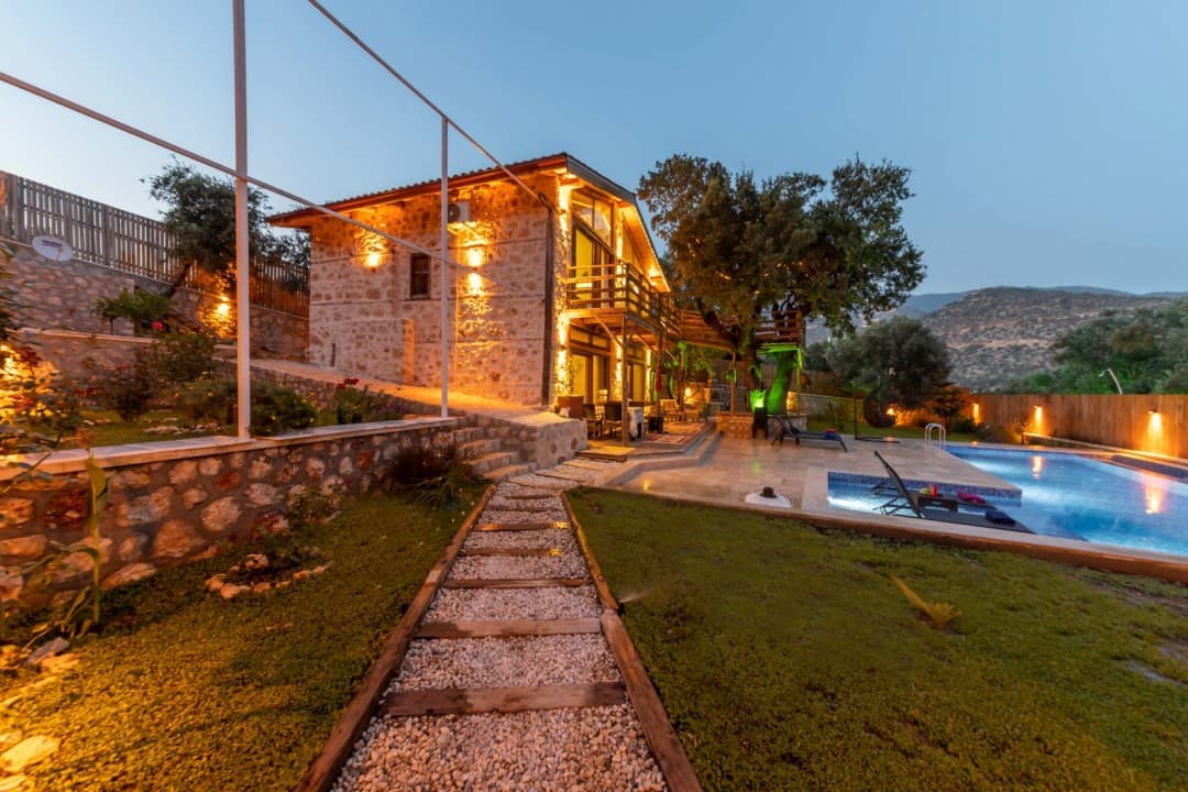 Taş döşeli yürüyüş yolu, havuz ve akşam ışıklandırılmış iki katlı taş villa.