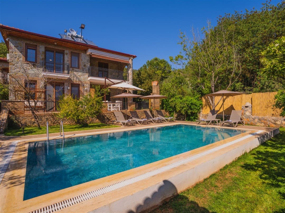 Taş mimarili iki katlı villa, önünde şezlonglu yüzme havuzu ve ağaçlarla çevrili bahçe.