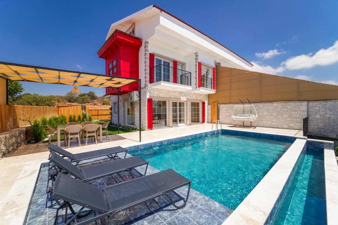 Kırmızı cumbalı iki katlı villa, önünde yüzme havuzu, şezlonglar ve gölgelikli yemek alanı.