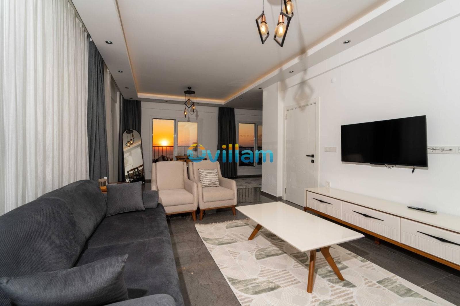 Gün batımı manzaralı pencereleri olan, gri koltuklu ve TV'li oturma odası.