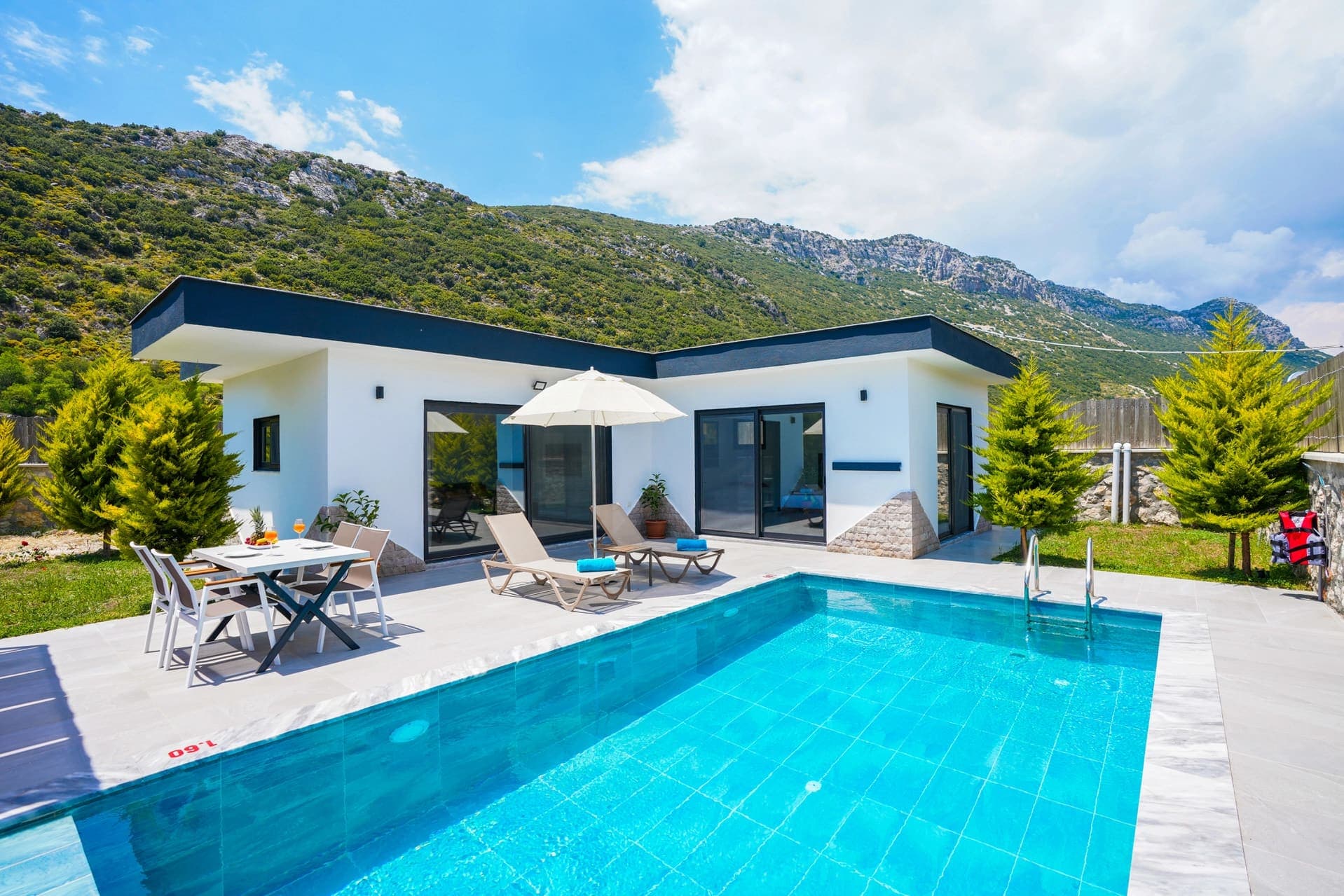 Dağ manzaralı havuz başında şezlonglar, yemek masası ve tek katlı villa.