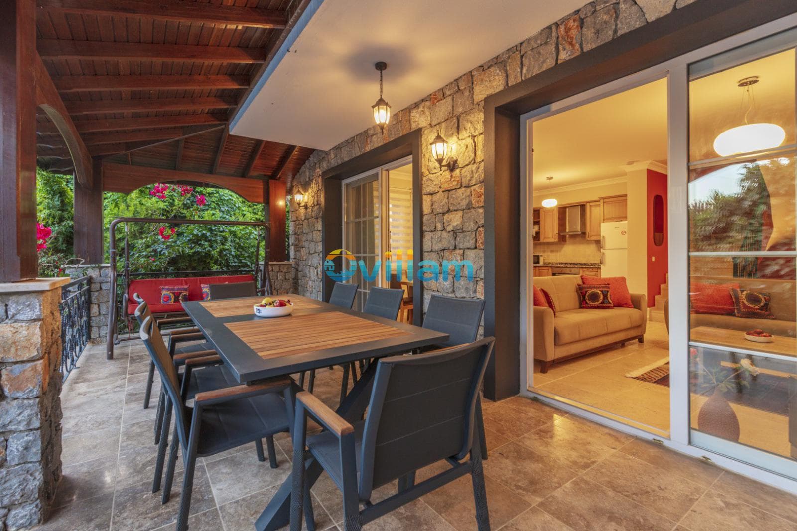 Taş duvarlı verandada yemek masası, sandalyeler ve salıncak.