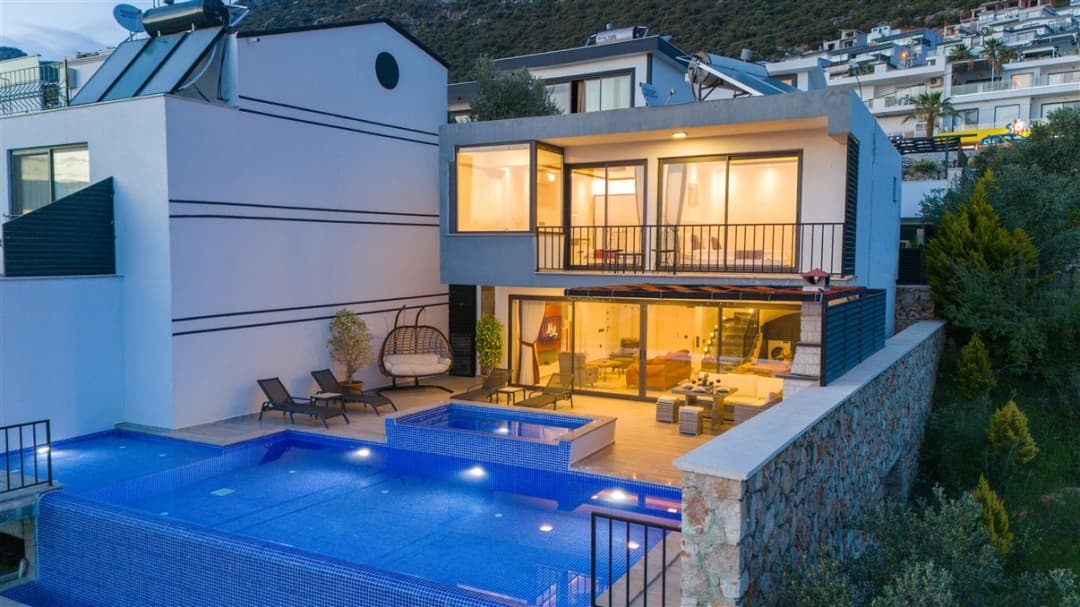 Işıkları yanan modern villa, önünde aydınlatılmış havuz, şezlonglar, salıncak ve taş duvarlı teras.
