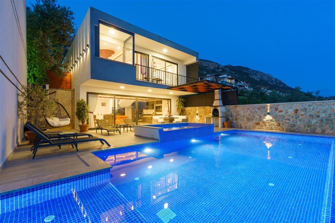 Gece aydınlatmalı havuzu, şezlongları ve barbeküsü olan modern villanın dış cephesi.