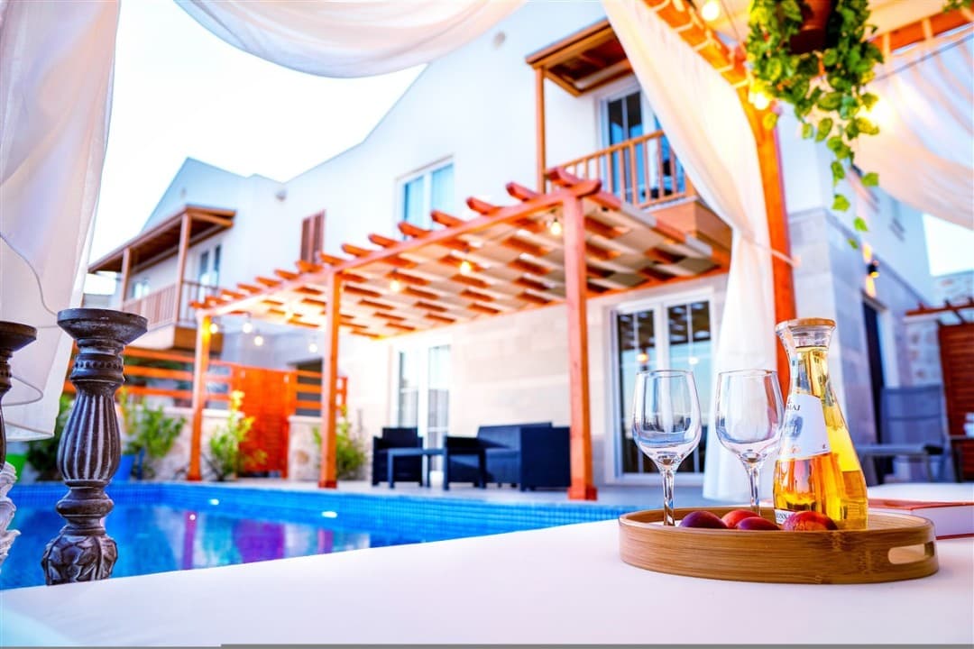 Ahşap pergola ve havuz kenarında şarap kadehleri bulunan beyaz villanın dış cephesi.