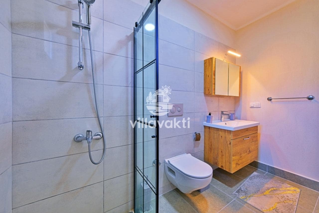 Cam duş kabini, duvara monte klozet ve ahşap lavabo dolabı olan modern banyo.